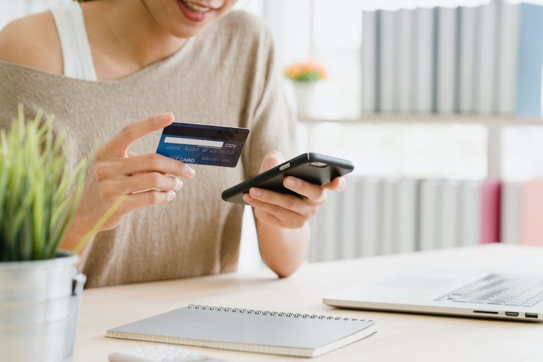 Beautiful Asian woman using smartphone buying online shopping by credit card while wear sweater sitting on desk in living room at home. Lifestyle woman at home concept